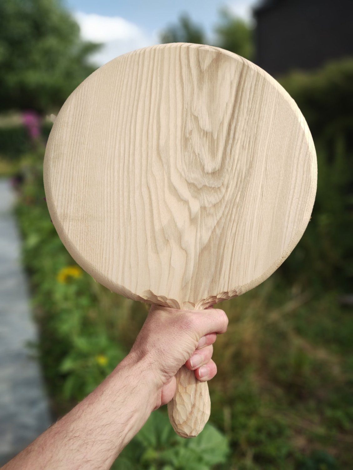 Round wooden pizza board held by hand outdoors, wood grain visible