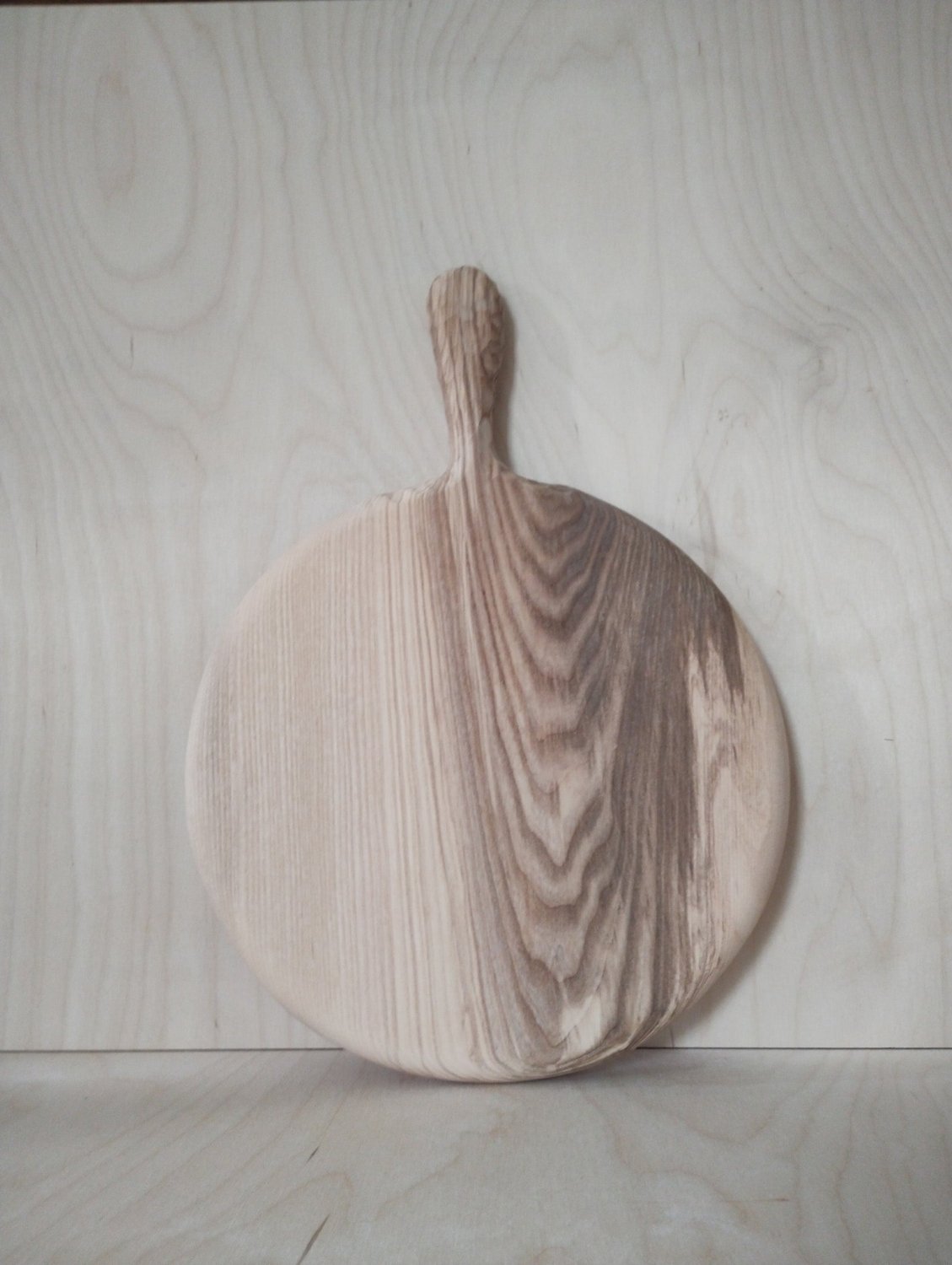 Round wooden pizza board with raised handle resting against light wood backdrop