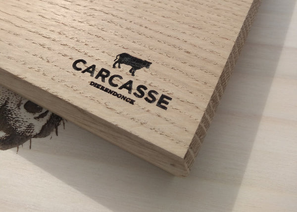 A beige wooden cutting board with a logo engraved on the side, placed on a wooden surface.